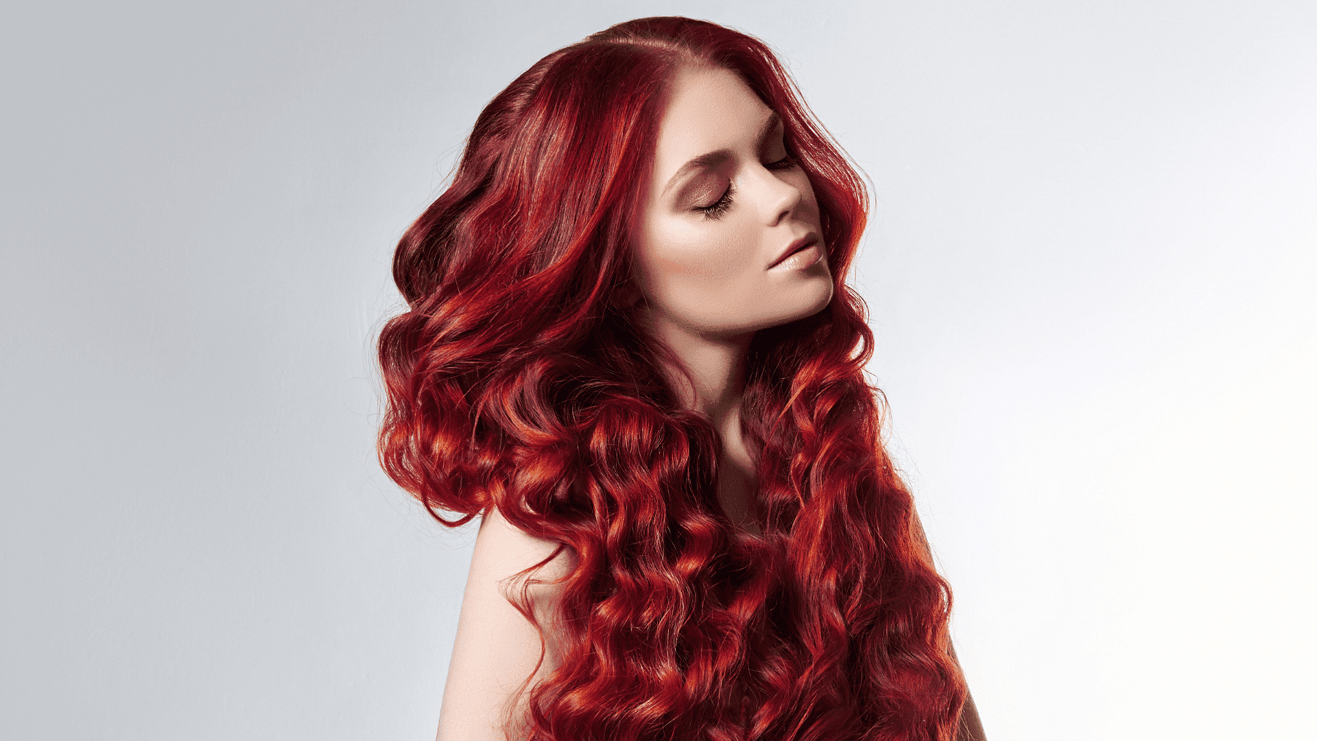 Woman with long, curly, vibrant red hair looking down against a plain background.