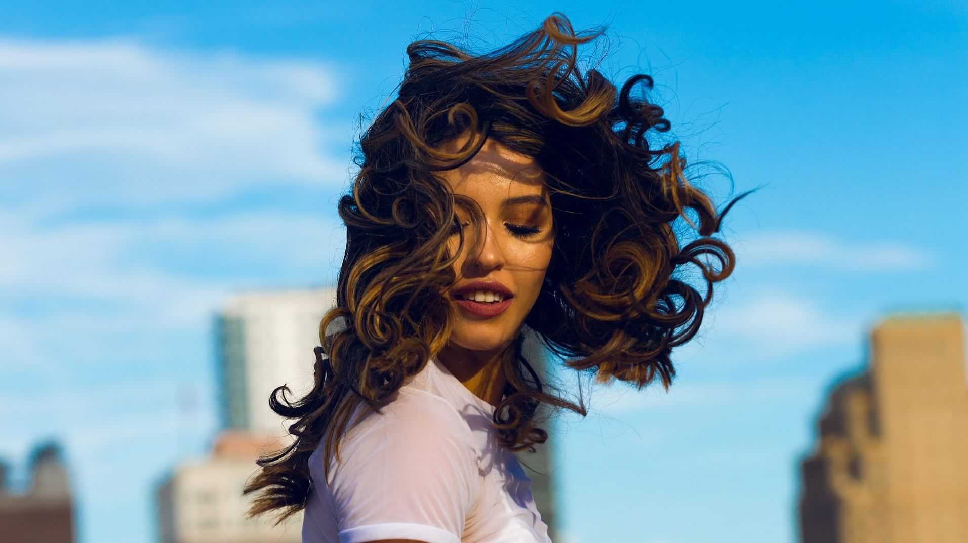 Woman with curly hair smiling against a cityscape and blue sky backdrop.